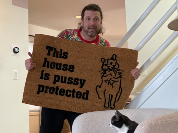 photo of a man showing a doormat reading This house is Pussy protected toa cat