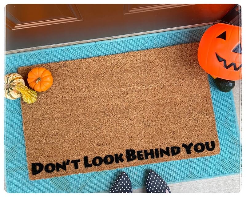 "A funny horror-themed Halloween doormat on a real decorated porch, October 2026."