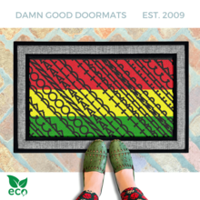 a pair of feet in black and white striped rainboots standing on an all-weather colorful, patterned all weather doormat with a geometric Fuck Donald Trump design in pan-African red, yellow, and green.