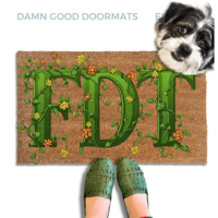 A dog standing on a coir doormat with the word "FDT" written on it in green vines, with a person's feet visible underneath.