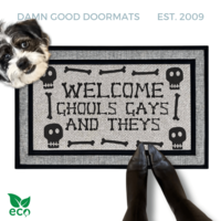 a humorous LGBTQ Halloween door mat with the text "Welcome Ghouls, Gays and Theys". There is also a terrier and a person's feet visible in the image.