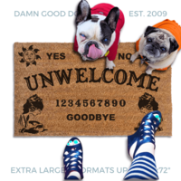 a welcome mat with a french bulldog and a fawn pug in bright hoodies, the word "UN-WELCOME" on it. The mat has a Ouija board design and the numbers "1234656789" printed on it. The scene includes a person's feet wearing white shoes, a vintage Halloween skull and cat light, and the text "DAMN GOOD DOORMATS EST. 2009" is visible.
