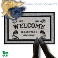 a black pug in a golden dragon wings costume standing on a welcome mat with the word "WELCOME" on it. The mat has a Ouija board design and the numbers "1234656789" printed on it. The scene includes a person's feet wearing black pointy toed boots and the text "DAMN GOOD DOORMATS EST. 2009" is visible.