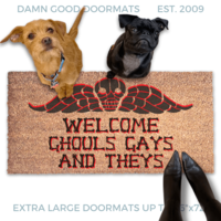 a humorous LGBTQ Halloween door mat with the text "Welcome Ghouls, Gays and Theys" and terrier and a black pug on top of it. There is also a pumpkin and a person's feet visible in the image.
