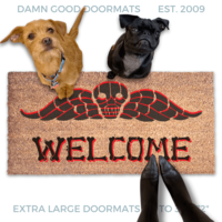 a dog and a small black pug and a terrier sitting on a welcome mat with a Beetlejuice skull design. The mat has the word "WELCOME" written on it in bones lettering