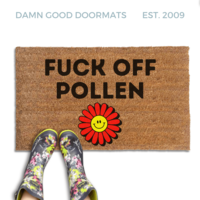 promotional image of a coir doormat reading "fuck off pollen" in with a red and yellow 70's hippy smiley faced flower on the bottom. A person in floral rain boots stands on the doormat