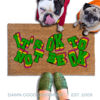 A pug and french bulldog in hoodies standing on a doormat with the text "IT'S OK TO BE OK" written on it, along with a person's feet wearing green shoes.