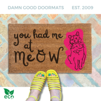 a pair of yellow strappy sandals on a brick floor, with a funny doormat. The text on the welcome mat reads "you had me at meow".