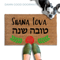 coir doormat reading Shana Tova in english and hebrew and a decorative pomegranate and wreath border.with a person in green slipons and a french bulldog