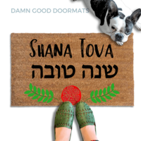 coir doormat reading Shana Tova in english and hebrew and a decorative pomegranate and wreath border.with a person in green slipons and a french bulldog