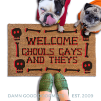 a humorous LGBTQ Halloween door mat with the text "Welcome Ghouls, Gays and Theys". There is also a pug and french bulldog in hoodies and a person's feet visible in the image.