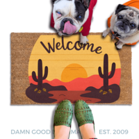 A pug and a french bulldog standing on a welcome mat with a desert sunset with cactus and landscape design, and a person's feet wearing green shoes in the foreground.