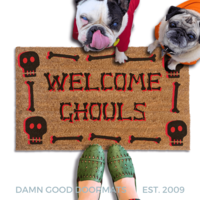 A french bulldog and a fawn pug in hoodies peeking over a welcome mat with the text "WELCOME GHOULS" and Halloween skull-themed decorations.