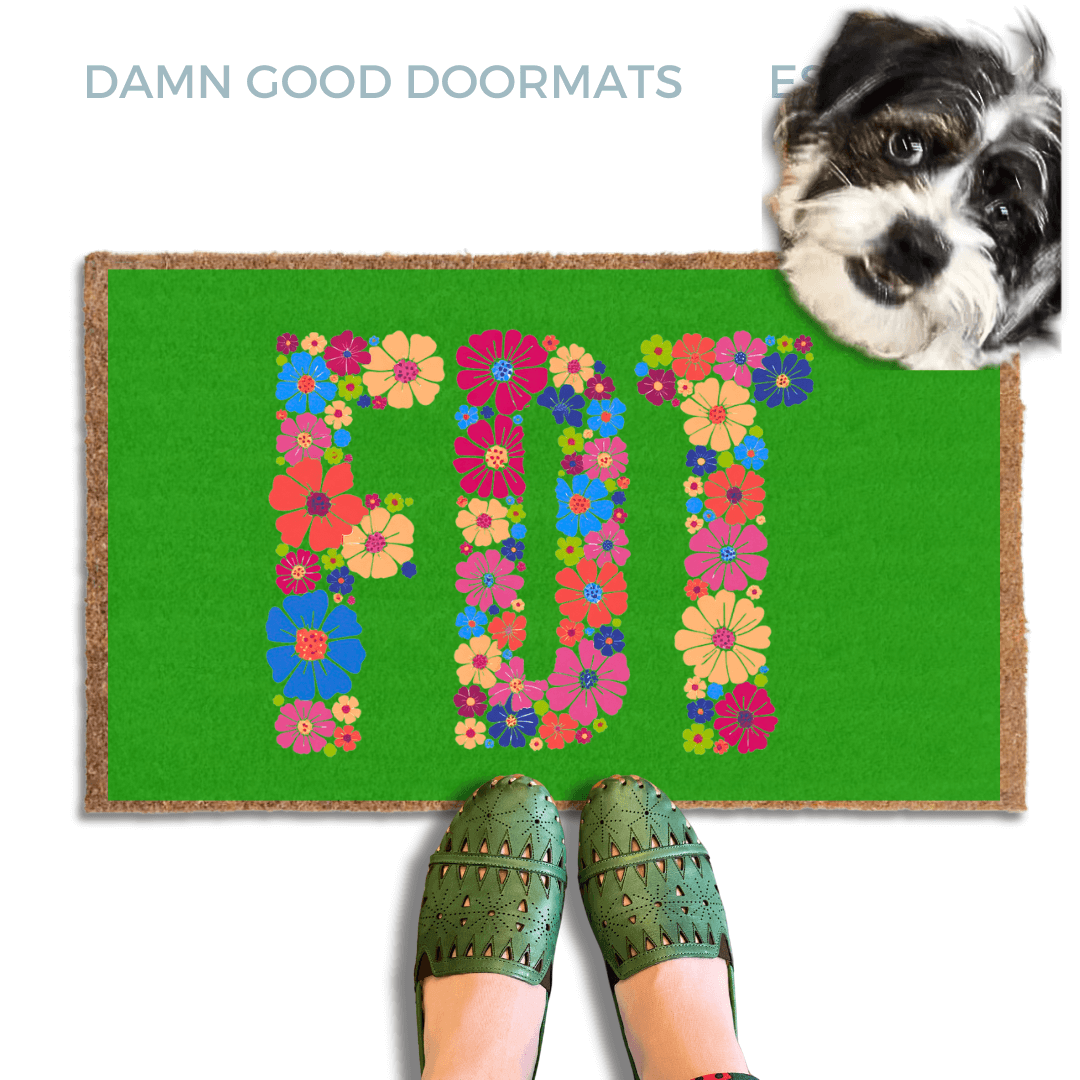 A terrier dog standing on a green doormat with the word "FDT" written on it, with a person's feet visible underneath.