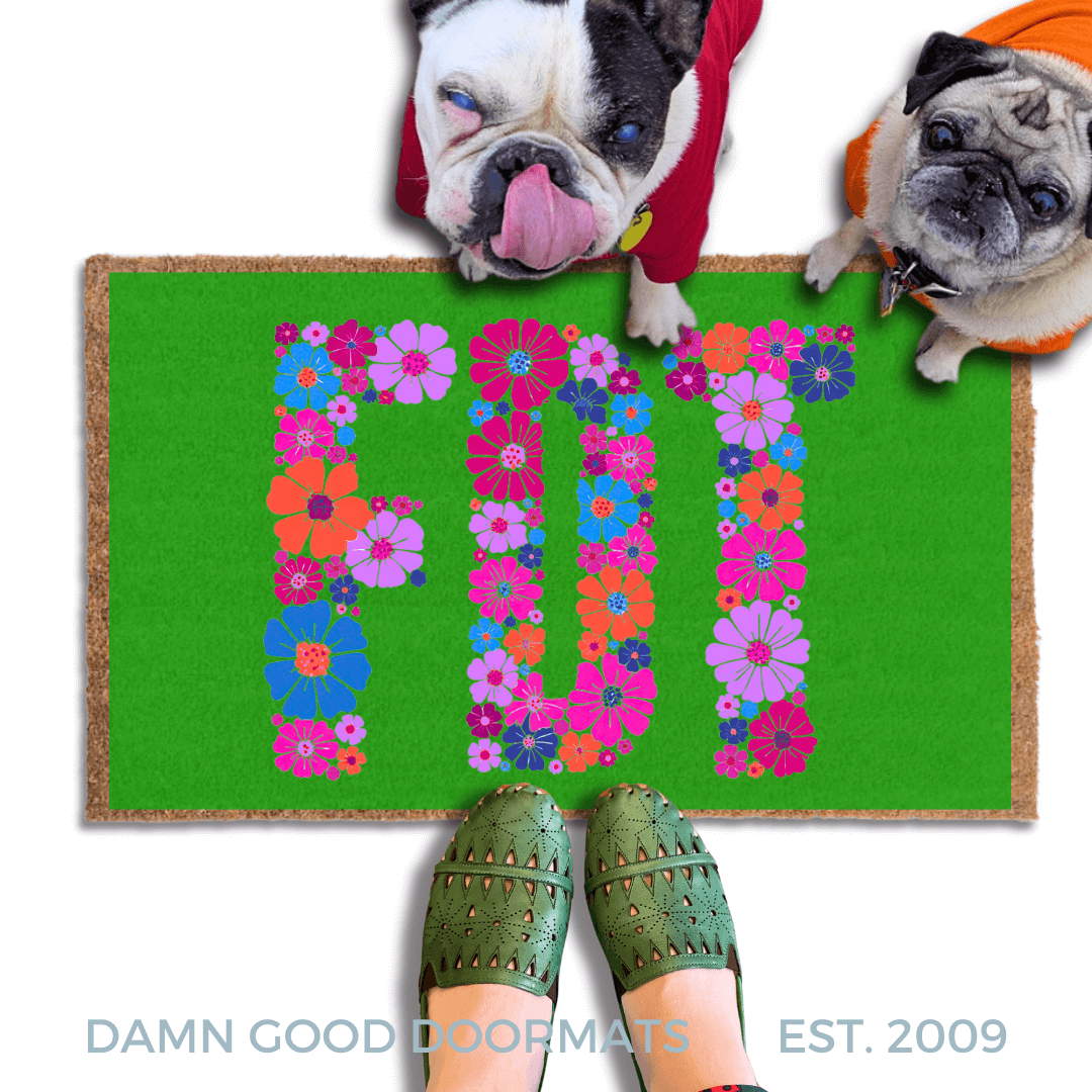 A pug and a french bulldog standing on a green doormat with the word "FDT" written on it in purple flowers, with a person's feet visible underneath.