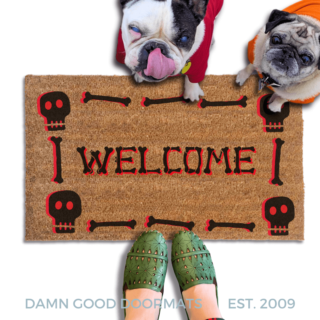 a Halloween-themed welcome mat with a skeleton and skull motif, a brindle french bulldog and a fawn pug in hoodies look up and a pair of feet in witchy boots stand on the doormat.