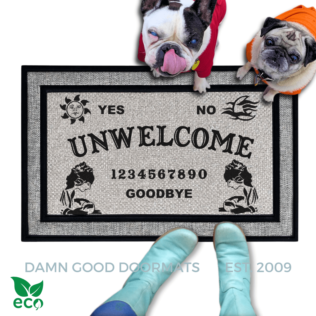 aa all weather gray doormat with a french bulldog and a fawn pug in bright hoodies, the word "UN-WELCOME" on it. The mat has a Ouija board design and the numbers "1234656789" printed on it. The scene includes a person's feet wearing white shoes, a vintage Halloween skull and cat light, and the text "DAMN GOOD DOORMATS EST. 2009" is visible.