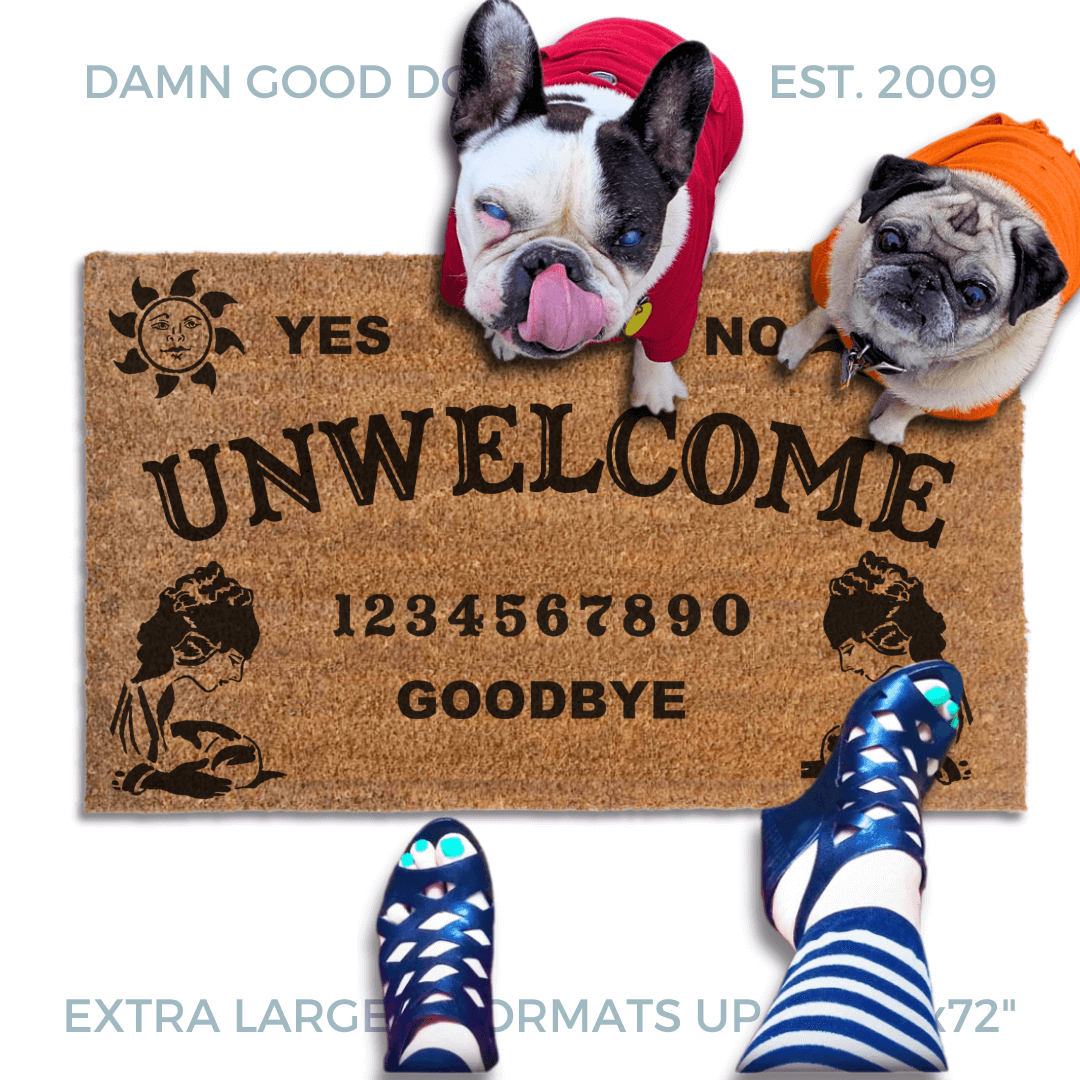 a welcome mat with a french bulldog and a fawn pug in bright hoodies, the word "UN-WELCOME" on it. The mat has a Ouija board design and the numbers "1234656789" printed on it. The scene includes a person's feet wearing white shoes, a vintage Halloween skull and cat light, and the text "DAMN GOOD DOORMATS EST. 2009" is visible.