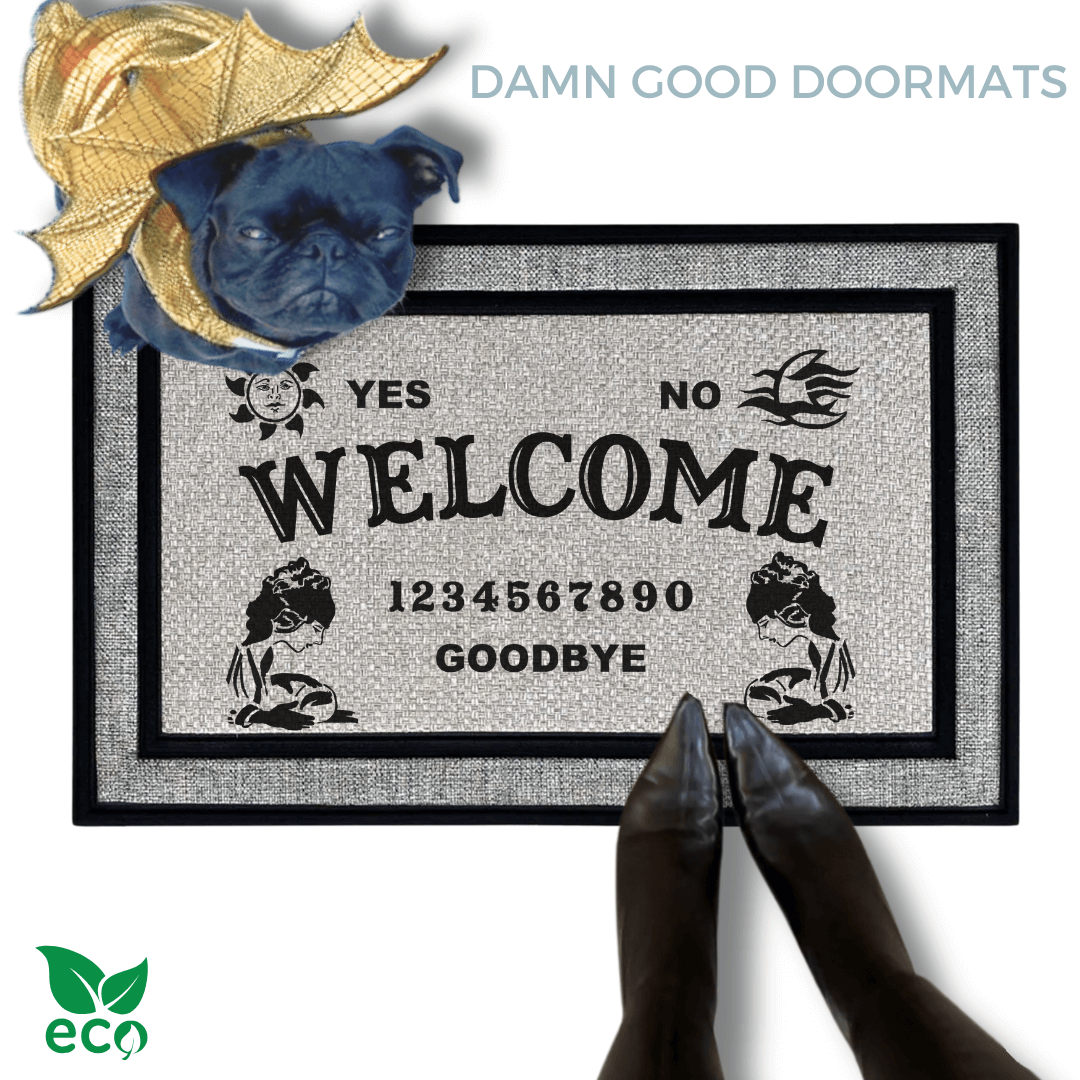 a black pug in a golden dragon wings costume standing on a welcome mat with the word "WELCOME" on it. The mat has a Ouija board design and the numbers "1234656789" printed on it. The scene includes a person's feet wearing black pointy toed boots and the text "DAMN GOOD DOORMATS EST. 2009" is visible.