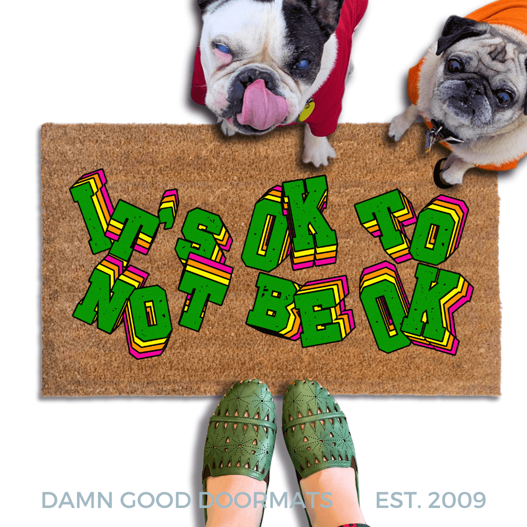A pug and french bulldog in hoodies standing on a doormat with the text "IT'S OK TO BE OK" written on it, along with a person's feet wearing green shoes.