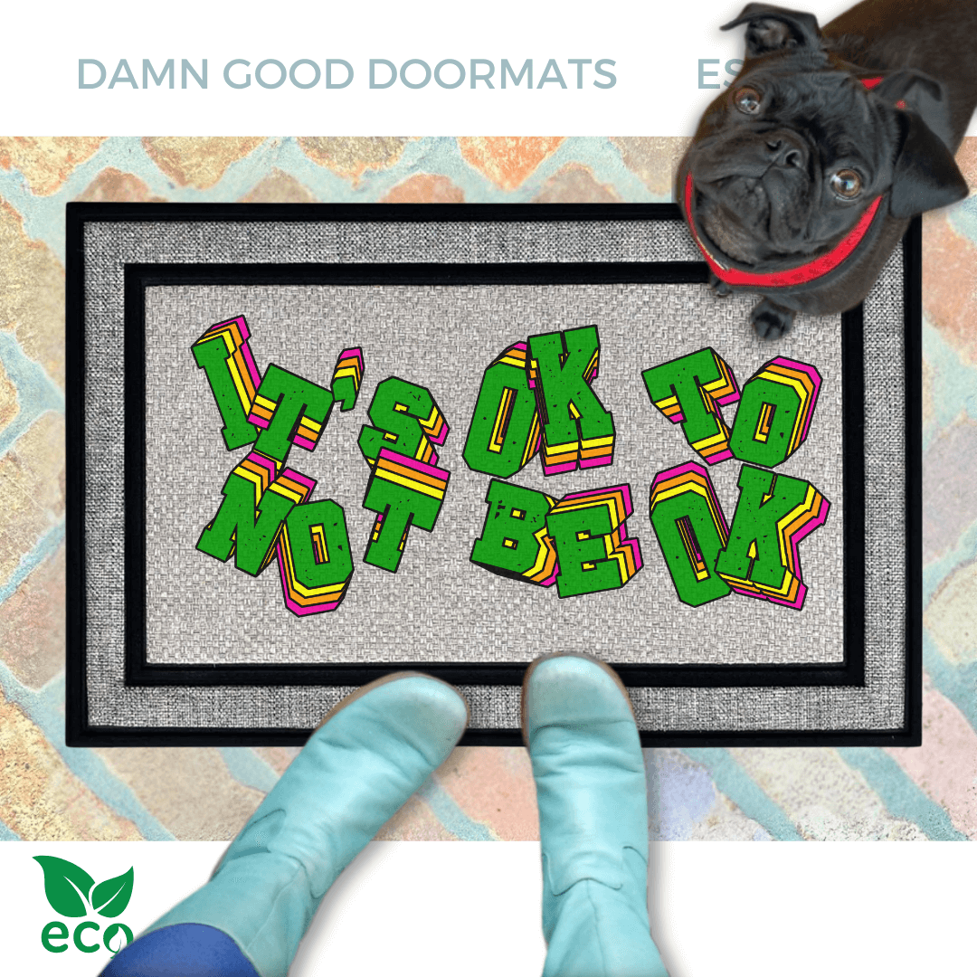 A black pug standing on an all weather doormat with the text "IT'S OK TO BE OK" written on it, along with a person's feet wearing smurf blue boots