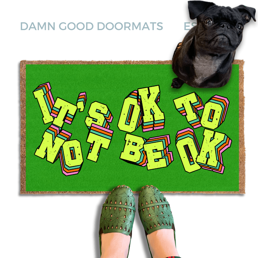 A pair of green shoes and a black pug dog are featured on a green doormat with the text "It's OK to BE OK" written on it.