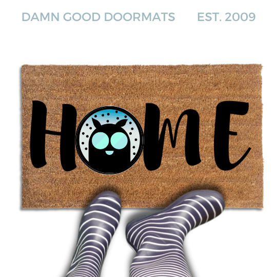 Coir doormat printed with HOME in black brush lettering, with Mothman's dark round silhouette and glowing teal eyes standing in for the O