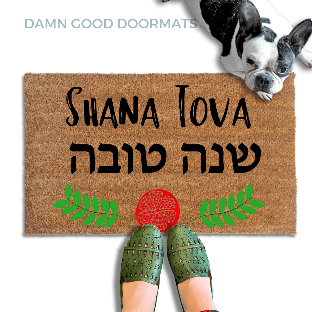 coir doormat reading Shana Tova in english and hebrew and a decorative pomegranate and wreath border.with a person in green slipons and a french bulldog
