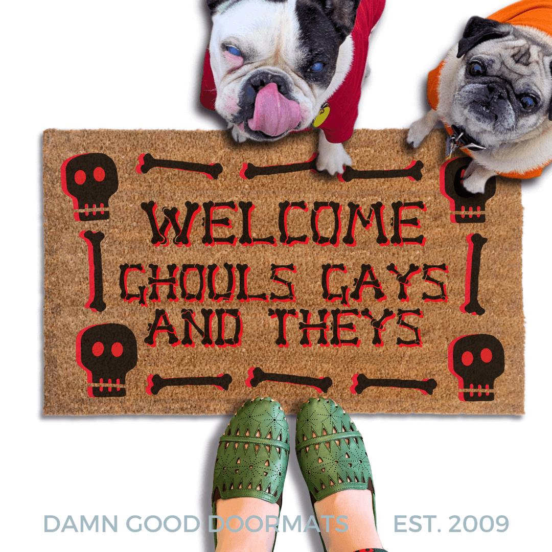 a humorous LGBTQ Halloween door mat with the text "Welcome Ghouls, Gays and Theys". There is also a pug and french bulldog in hoodies and a person's feet visible in the image.