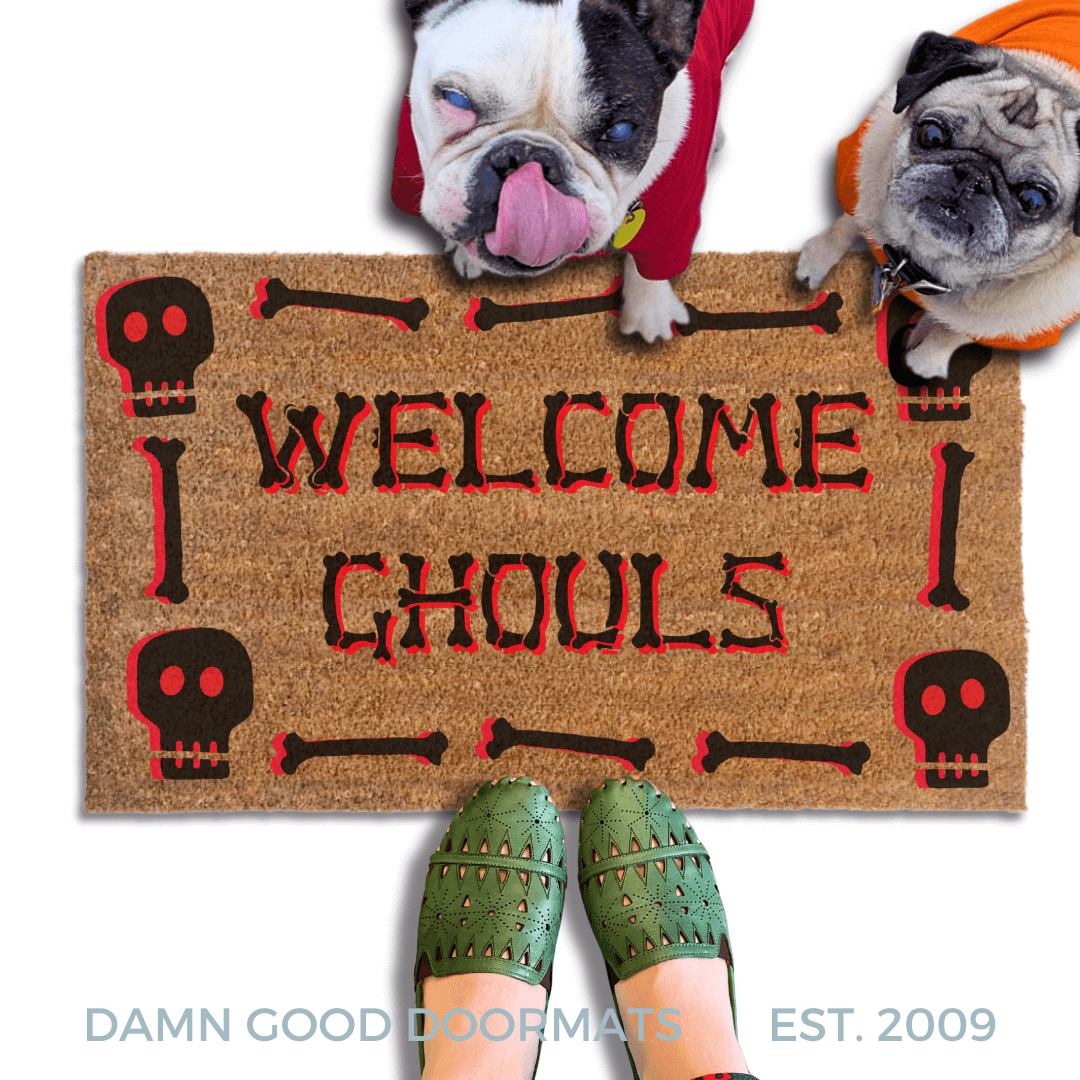 A french bulldog and a fawn pug in hoodies peeking over a welcome mat with the text "WELCOME GHOULS" and Halloween skull-themed decorations.