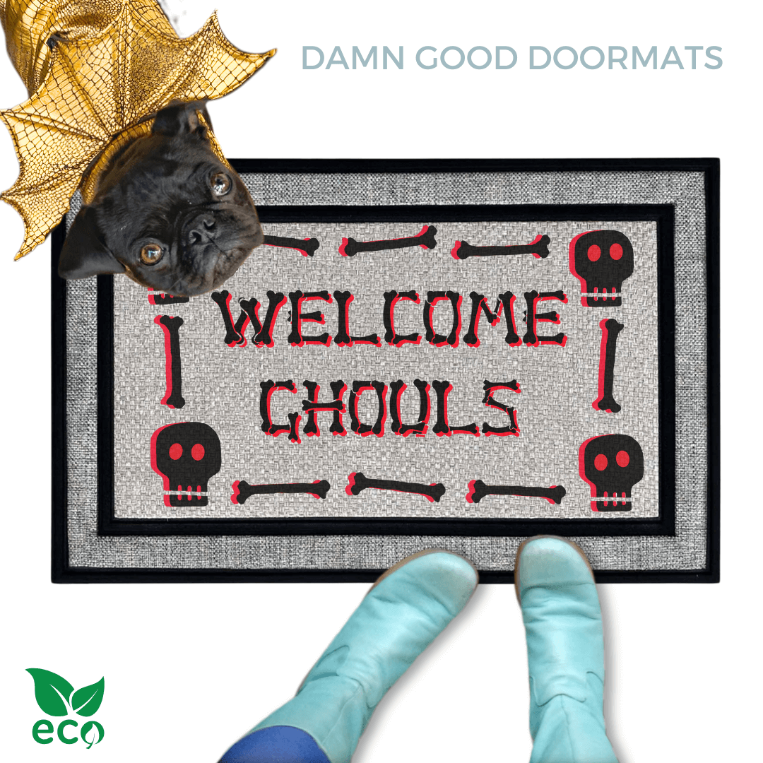 A black pug in gold dragon wings halloween costume peeking over a welcome mat with the text "WELCOME GHOULS" and Halloween skull-themed decorations with a person in smurf blue boots.
