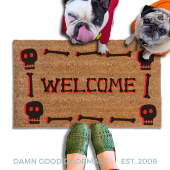 a Halloween-themed welcome mat with a skeleton and skull motif, a brindle french bulldog and a fawn pug in hoodies look up and a pair of feet in witchy boots stand on the doormat.