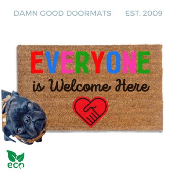 Everyone is welcome here doormat with a black pug standing on it looking up