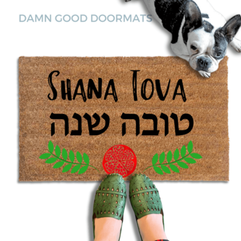 coir doormat reading Shana Tova in english and hebrew and a decorative pomegranate and wreath border.with a person in green slipons and a french bulldog