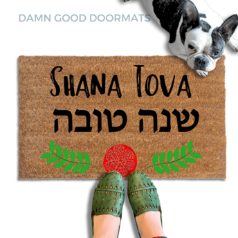 coir doormat reading Shana Tova in english and hebrew and a decorative pomegranate and wreath border.with a person in green slipons and a french bulldog
