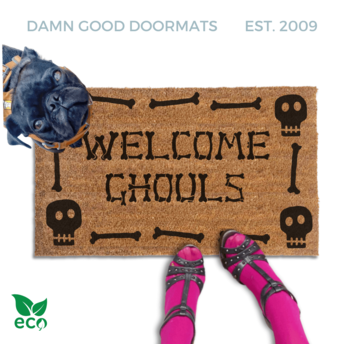 a dog standing next to a welcome mat with the text "WELCOME GHOULS" on it. The dog is wearing a collar and appears to be looking at the mat. The mat has a skull design. The image also includes a pair of feet wearing pink tights and high heels.