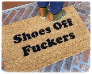 Natural coir doormat with bold black letters reading SHOES, FUCKERS, shown on a covered porch.