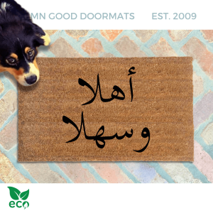 a dog peeking over a brown doormat with Arabic script on it reading welcome on two lines