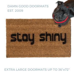 Natural coir doormat with black lettering reading Stay Shiny, Firefly, pictured with a black pug in a red harness looking up. Nerdy sci-fi welcome mat.