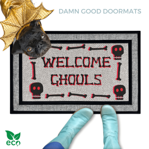 A black pug in gold dragon wings halloween costume peeking over a welcome mat with the text "WELCOME GHOULS" and Halloween skull-themed decorations with a person in smurf blue boots.