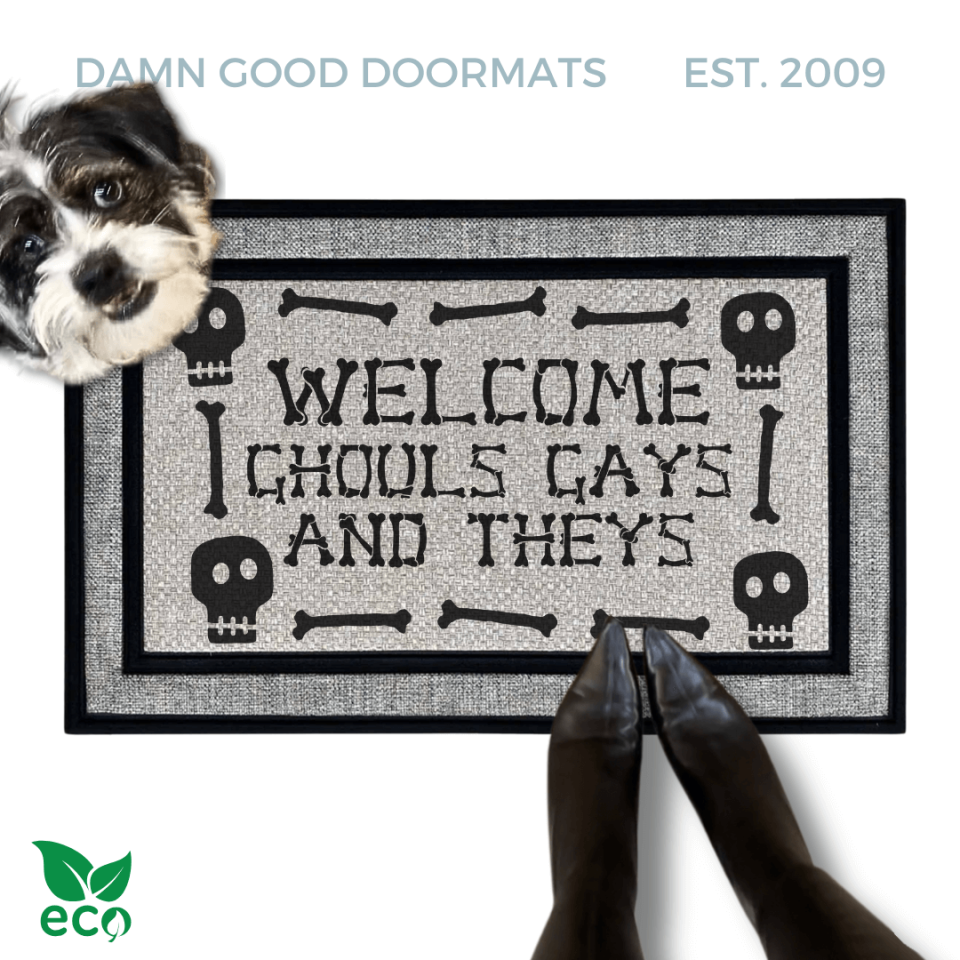 a humorous LGBTQ Halloween door mat with the text "Welcome Ghouls, Gays and Theys". There is also a terrier and a person's feet visible in the image.