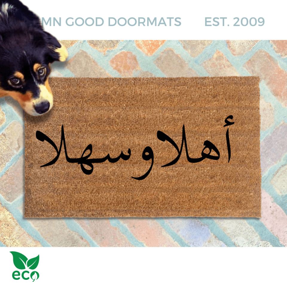 a dog peeking over a brown doormat with Arabic script on it reading welcome