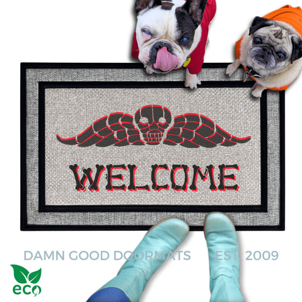 a dog and a fawn pug and a french bulldog in hoodies sitting on an all weather welcome mat with a Beetlejuice skull design. The mat has the word "WELCOME" written on it in bones lettering