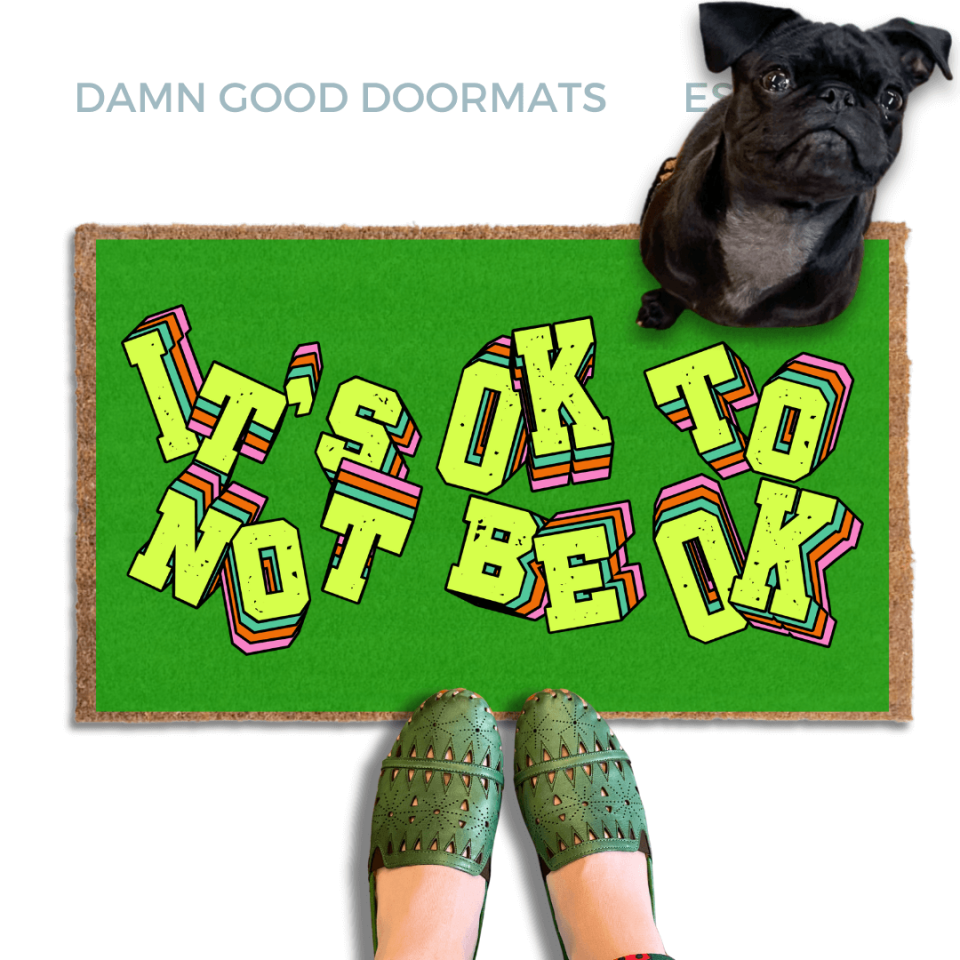 A pair of green shoes and a black pug dog are featured on a green doormat with the text "It's OK to BE OK" written on it.