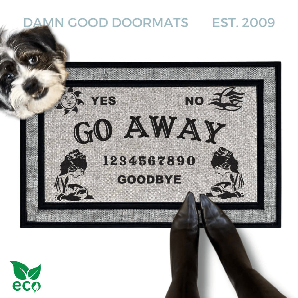 a gray all weather welcome mat with a terrier dog, the word "GO AWAY" on it. The mat has a Ouija board design and the numbers "1234656789" printed on it. The scene includes a person's feet wearing black pointy toed witch boots and the text "DAMN GOOD DOORMATS EST. 2009" is visible.