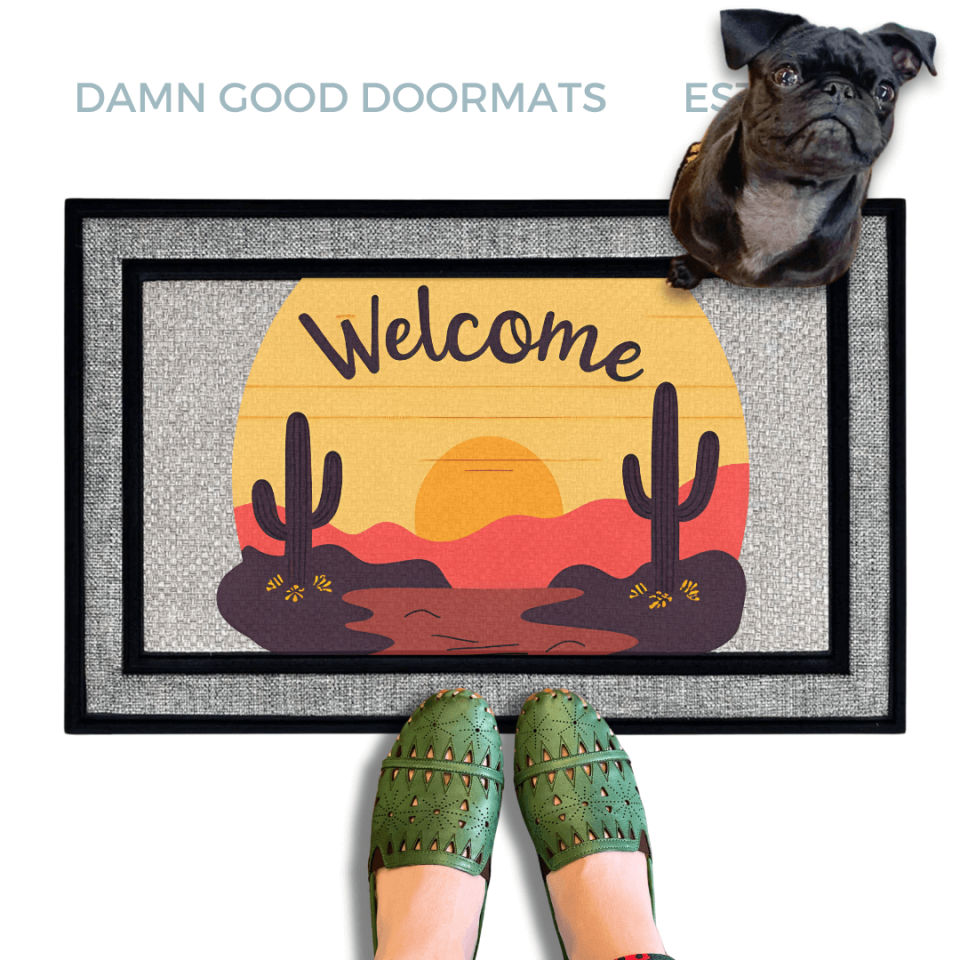 A pug standing on an all weather welcome mat with a desert sunset with cactus and landscape design, and a person's feet wearing green shoes in the foreground.