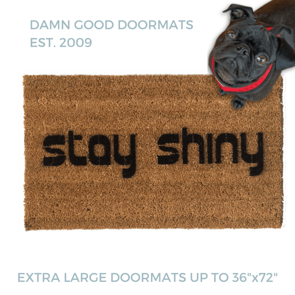 Natural coir doormat with black lettering reading Stay Shiny, Firefly, pictured with a black pug in a red harness looking up. Nerdy sci-fi welcome mat.