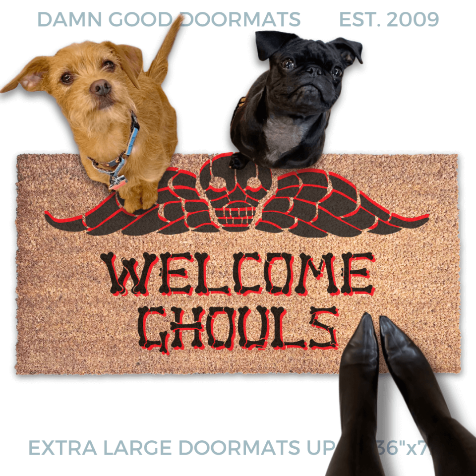 a terrier and a black pug dog sitting on a door mat with the text "WELCOME GHOULS" and a spooky Beetlejuice tombstone on it.