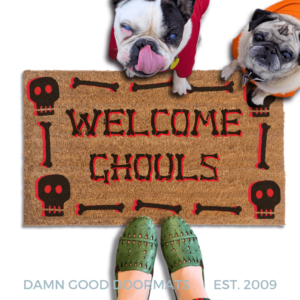 A french bulldog and a fawn pug in hoodies peeking over a welcome mat with the text "WELCOME GHOULS" and Halloween skull-themed decorations.