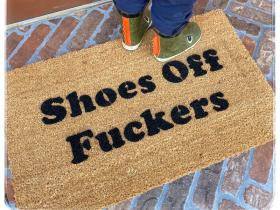 Natural coir doormat with bold black letters reading SHOES, FUCKERS, shown on a covered porch.